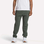 A person wearing a white t-shirt, Reebok Men's Patent Pants in slim fit olive green with an adjustable waistband, and white sneakers stands against a plain light background; their face is not visible.