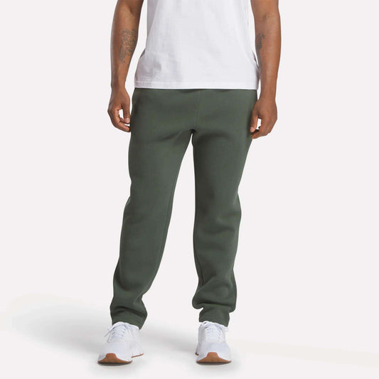 A person wearing a white t-shirt, Reebok Men's Patent Pants in slim fit olive green with an adjustable waistband, and white sneakers stands against a plain light background; their face is not visible.
