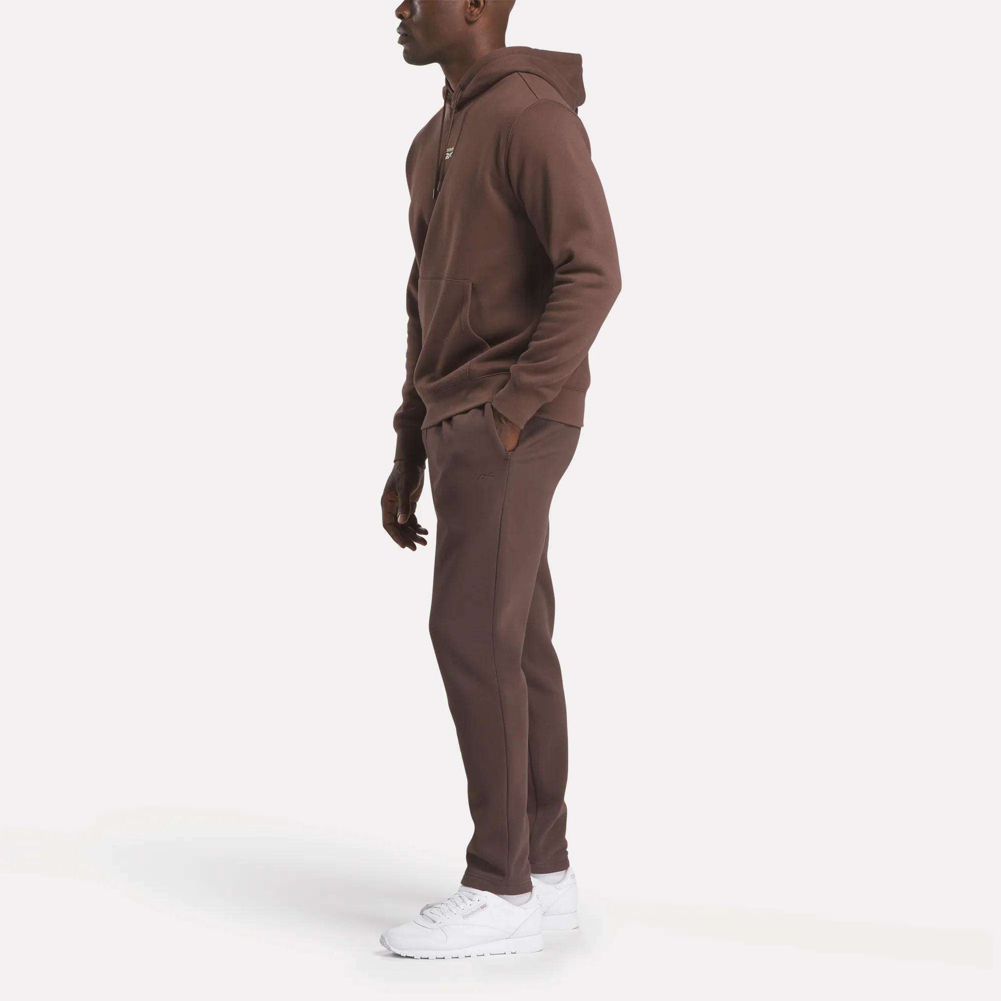 A person stands sideways in a Reebok Men's Finisher Hoodie and matching sweatpants with white sneakers, set against a plain white background.