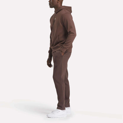 A person stands sideways in a Reebok Men's Finisher Hoodie and matching sweatpants with white sneakers, set against a plain white background.