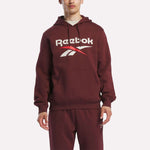 A person stands facing forward in a maroon Reebok Men's Logo Hoodie, featuring a bold chest logo and made from soft cotton-blend fleece for comfort. Matching sweatpants complete the look.