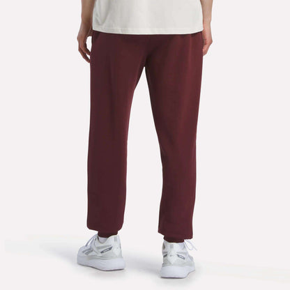 A person stands facing away against a plain light backdrop, showing only their lower half in maroon Reebok Men's French Terry Logo Joggers, paired with a white shirt and white sneakers.