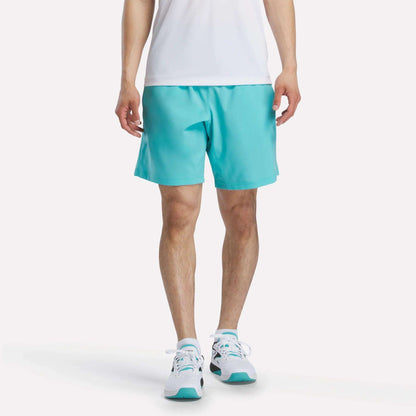 A person, head not visible, stands against a plain white background wearing a white shirt, Reebok Men's Woven Shorts in turquoise, and white sneakers with turquoise accents.