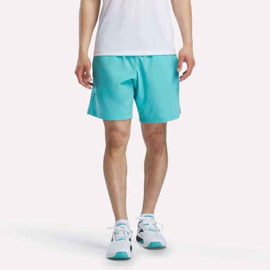 A person, head not visible, stands against a plain white background wearing a white shirt, Reebok Men's Woven Shorts in turquoise, and white sneakers with turquoise accents.
