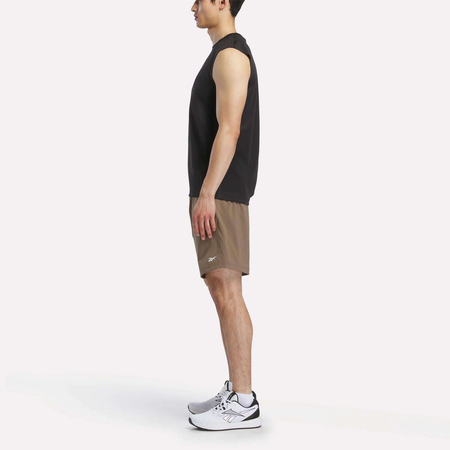 A man in profile wears a black sleeveless shirt, Reebok Men's Woven Shorts, and white athletic shoes against a plain white backdrop.