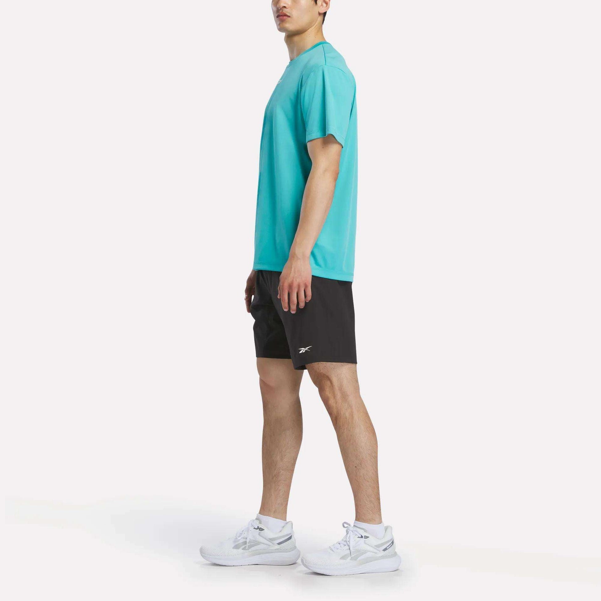 A man stands sideways in a Reebok Men's Short Sleeve Tech Training T-Shirt (turquoise), black Reebok shorts, and white sneakers against a plain white background. The gym shirt features sweat-wicking fabric for optimal comfort.