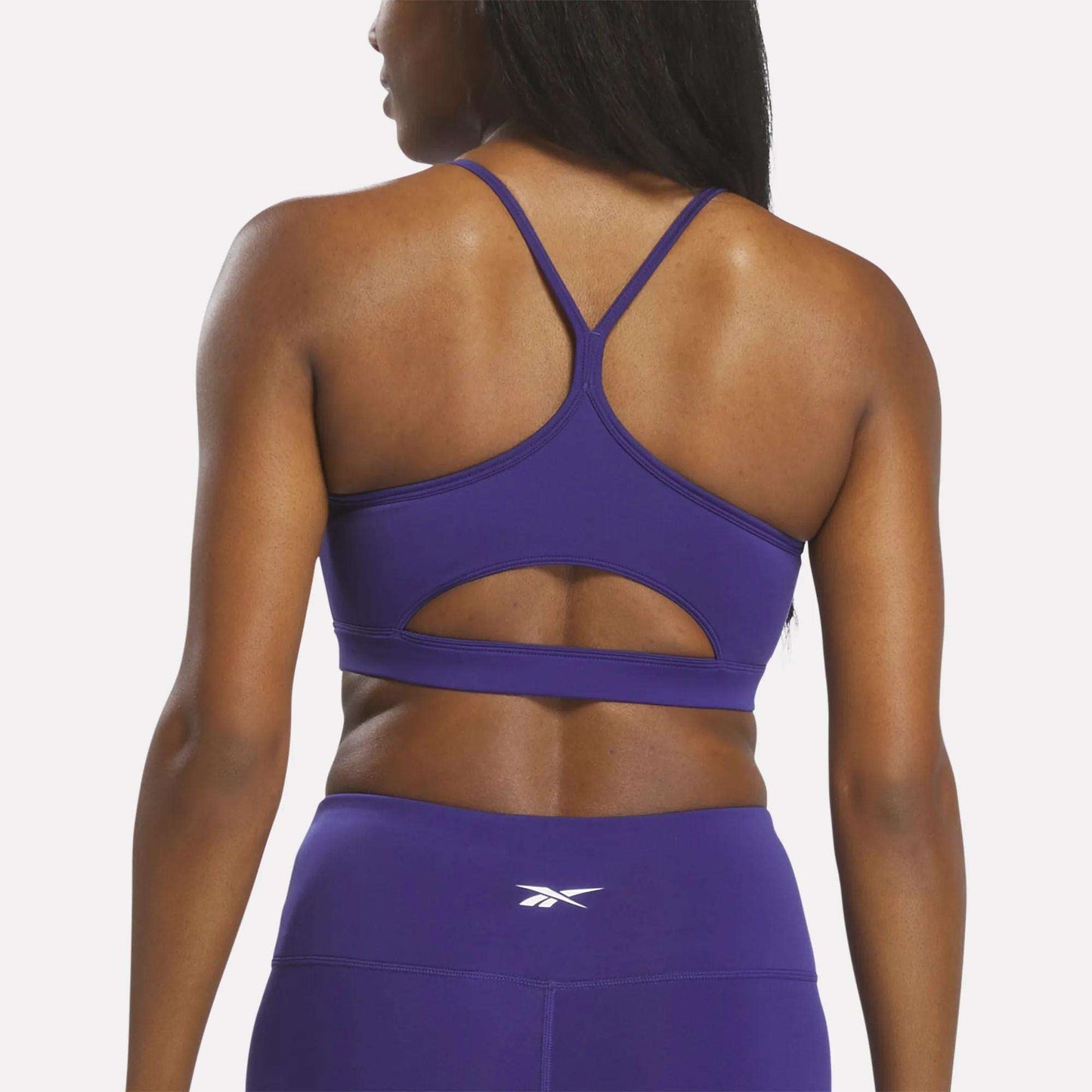 A woman is shown from the back against a plain white background, wearing the Reebok Women's Strappy Sports Bra in purple, featuring cutout detailing for medium-impact workouts and quick-drying comfort. She has paired it with matching high-waisted leggings.