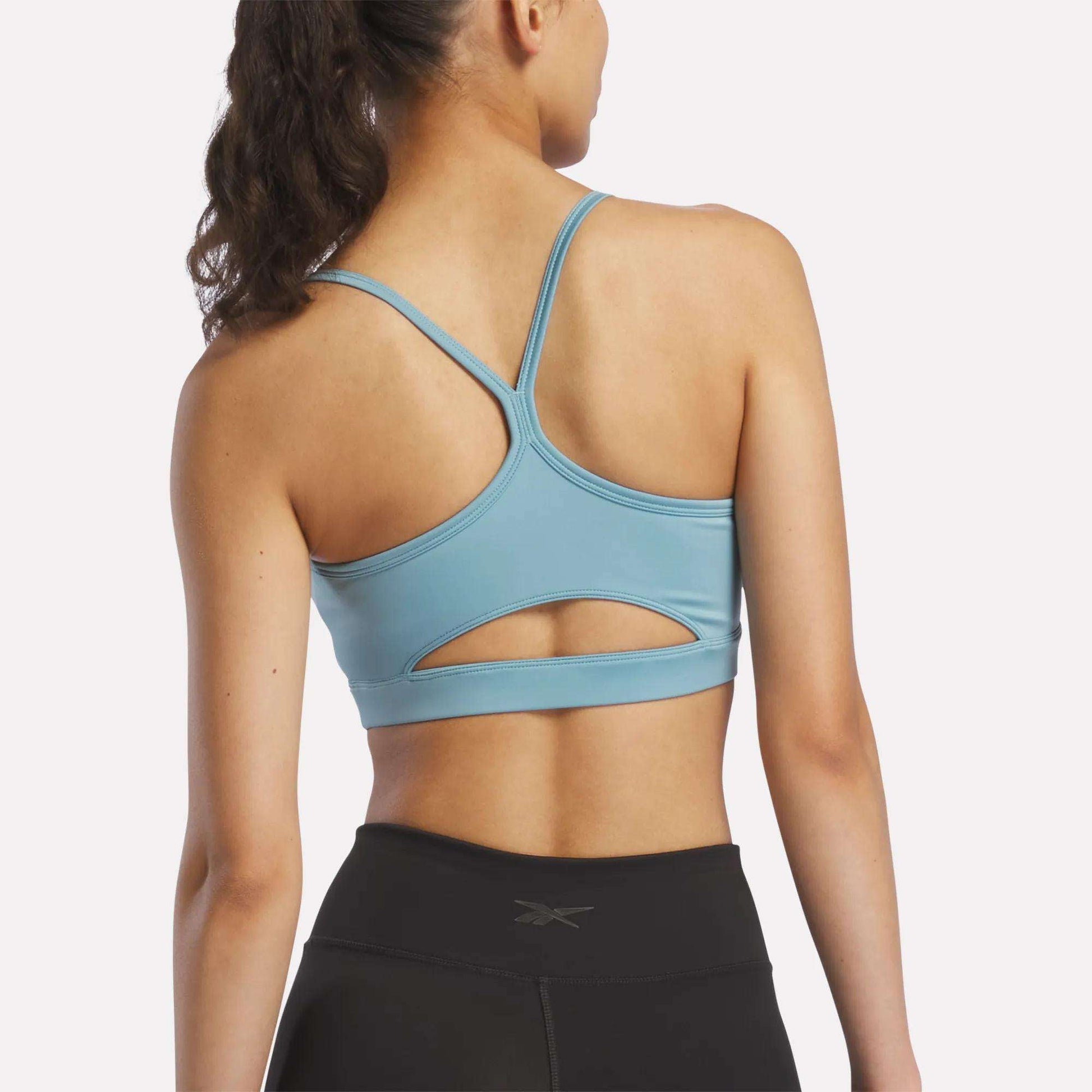 A woman with a ponytail is seen from behind against a plain background, wearing the Reebok Women's Strappy Sports Bra in light blue and black leggings. This quick-drying bra delivers support and comfort, making it perfect for medium-impact workouts.