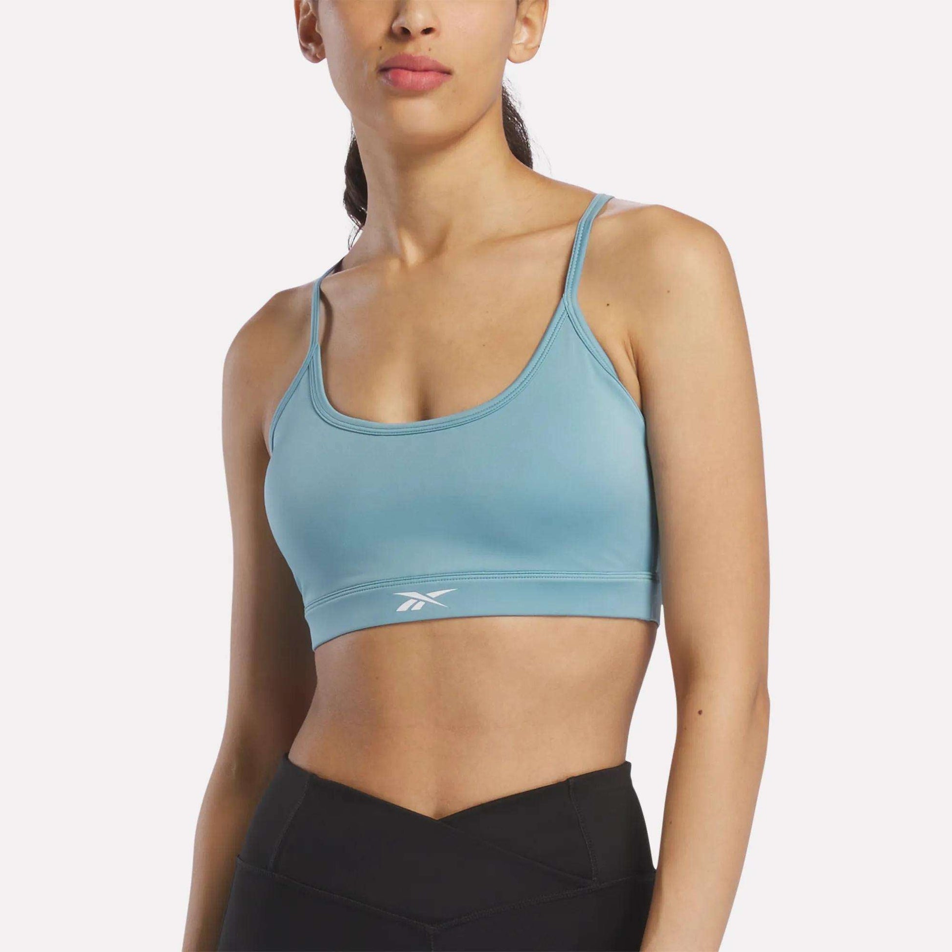 A woman wearing the Reebok Women's Strappy Sports Bra in Hazy Teal, ideal for medium-impact workouts thanks to its quick-drying fabric, and black leggings stands against a plain background.