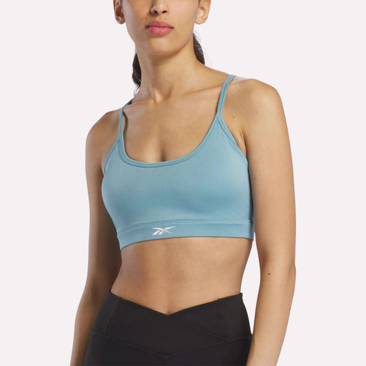 A woman wearing the Reebok Women's Strappy Sports Bra in Hazy Teal, ideal for medium-impact workouts thanks to its quick-drying fabric, and black leggings stands against a plain background.