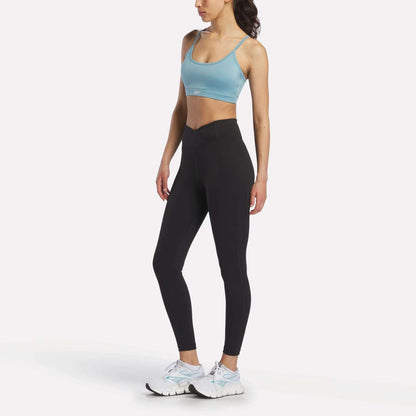 A woman stands against a plain white background, wearing the Reebok Women's Strappy Sports Bra in light blue—ideal for medium-impact workouts thanks to its quick-drying fabric—paired with black high-waisted leggings and white athletic shoes.
