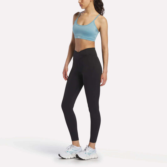 A woman stands against a plain white background, wearing the Reebok Women's Strappy Sports Bra in light blue—ideal for medium-impact workouts thanks to its quick-drying fabric—paired with black high-waisted leggings and white athletic shoes.