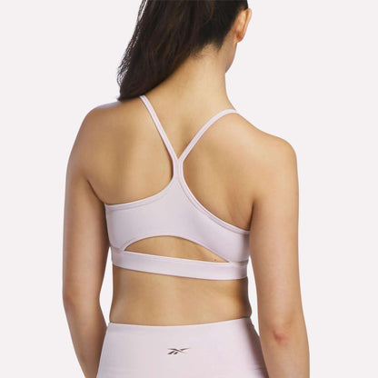 A woman with long dark hair is shown from the back against a plain white background, wearing the Reebok Women's Strappy Sports Bra in light pink, paired with matching leggings and designed for medium-impact workouts.