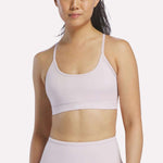 A woman wearing the Reebok Women's Strappy Sports Bra in Elemental Pink stands against a plain white background, ready for medium-impact workouts with the benefit of quick-drying fabric.