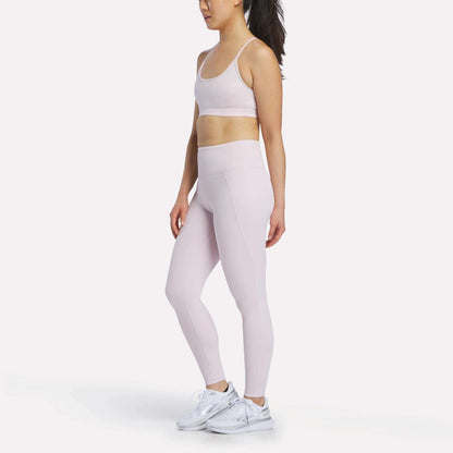 A woman stands sideways against a plain white background, wearing a light pink Reebok Women's Strappy Sports Bra and matching leggings with white sneakers. The quick-drying fabric of the sports bra is perfect for medium-impact workouts.