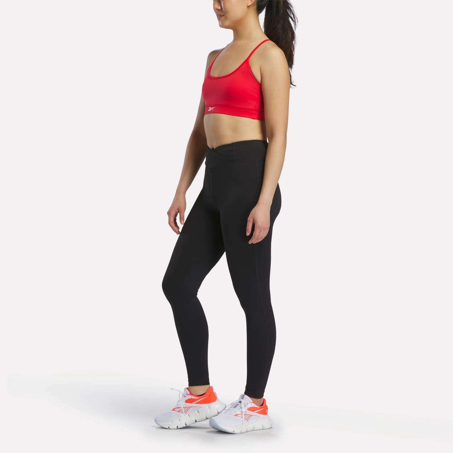 A woman stands sideways against a plain white background, wearing the Reebok Women's Strappy Sports Bra in red—ideal for medium-impact workouts—paired with black leggings and white athletic shoes featuring orange accents.