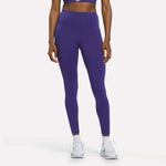A person wearing Reebok Women's High-Rise Leggings in Bold Purple, paired with a matching sports bra and white sneakers, stands against a plain white background. Their arms are relaxed by their sides, and their face is not visible.