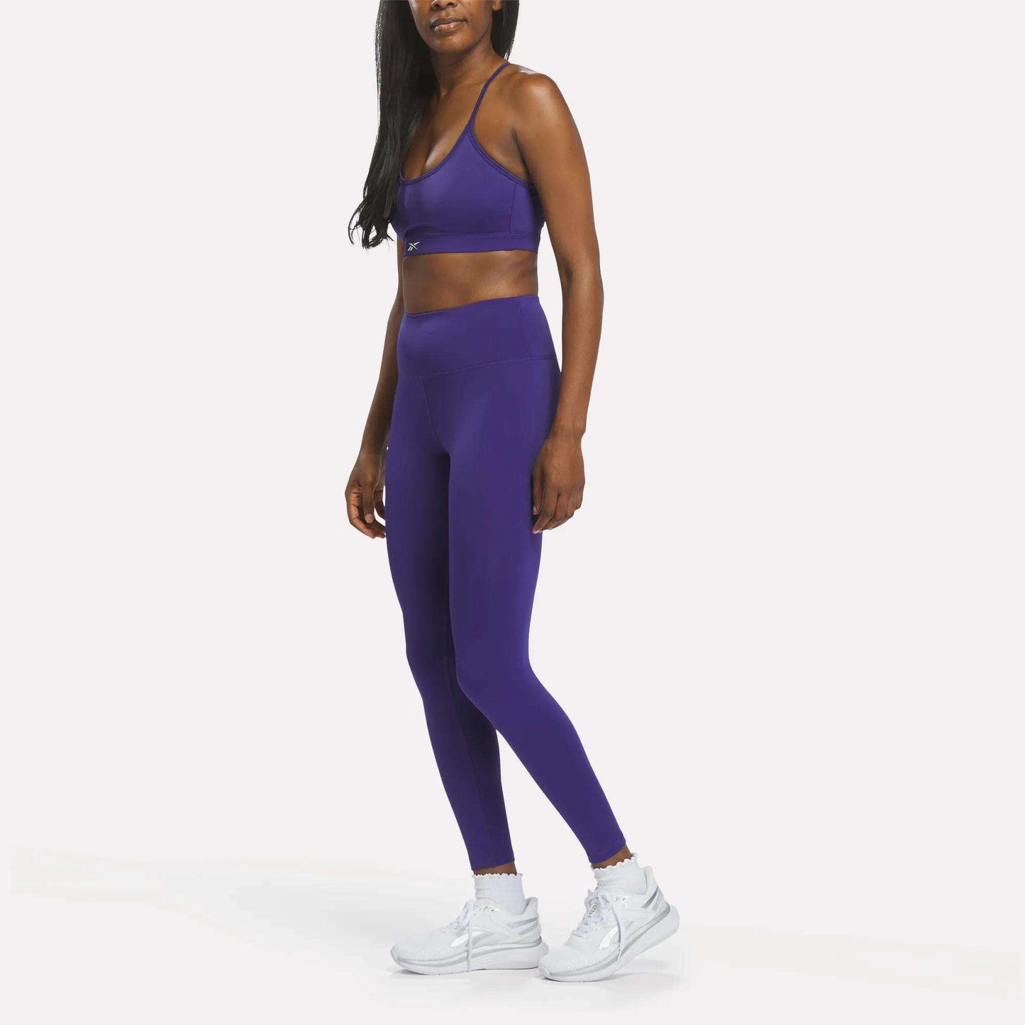 A woman stands against a plain white background wearing Reebok Women's High-Rise Leggings in purple, crafted from RBK-DRY fabric, paired with a matching sports bra and white sneakers.