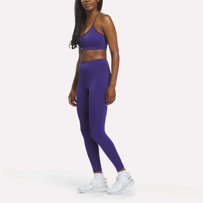 A woman stands against a plain white background wearing Reebok Women's High-Rise Leggings in purple, crafted from RBK-DRY fabric, paired with a matching sports bra and white sneakers.