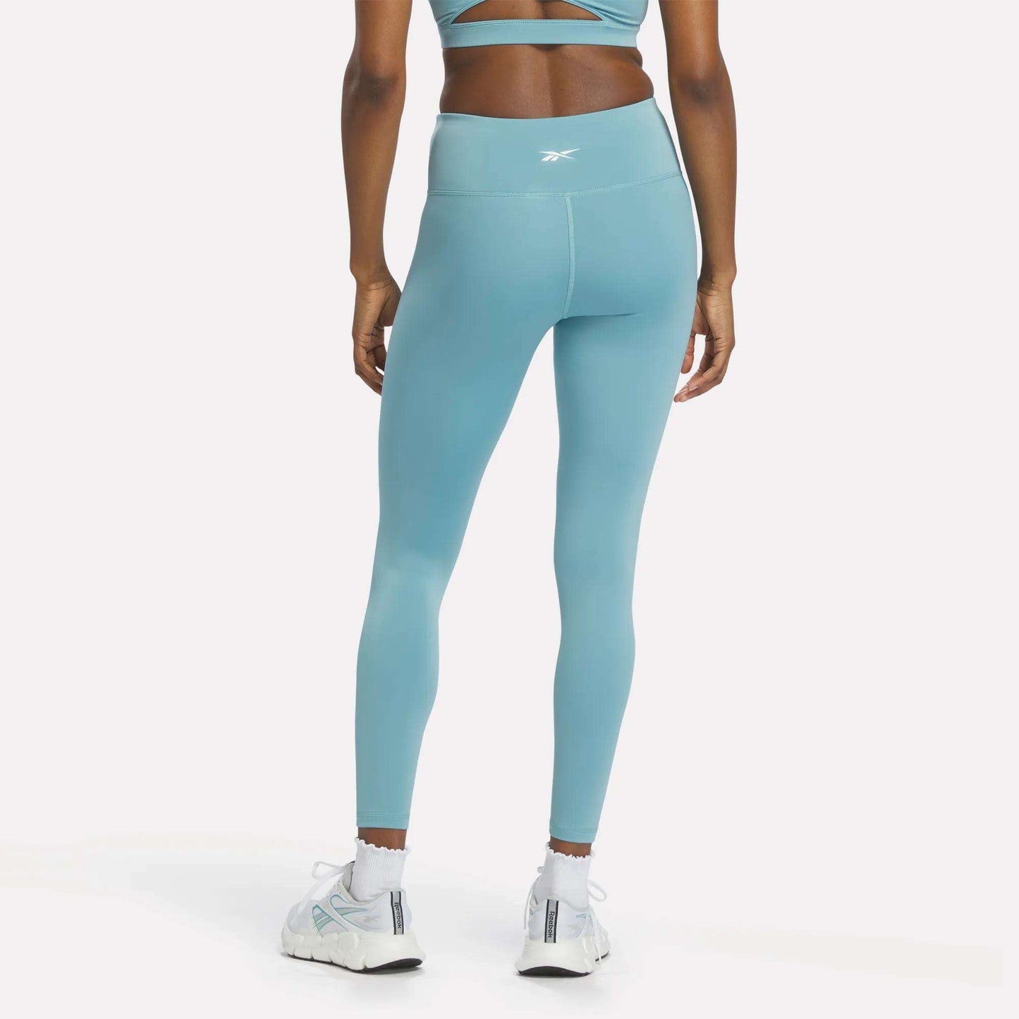 A person is seen from the back, standing against a plain white background, wearing Reebok Women's High-Rise Leggings in light blue made with RBK-DRY fabric, paired with a matching sports bra and white sneakers.