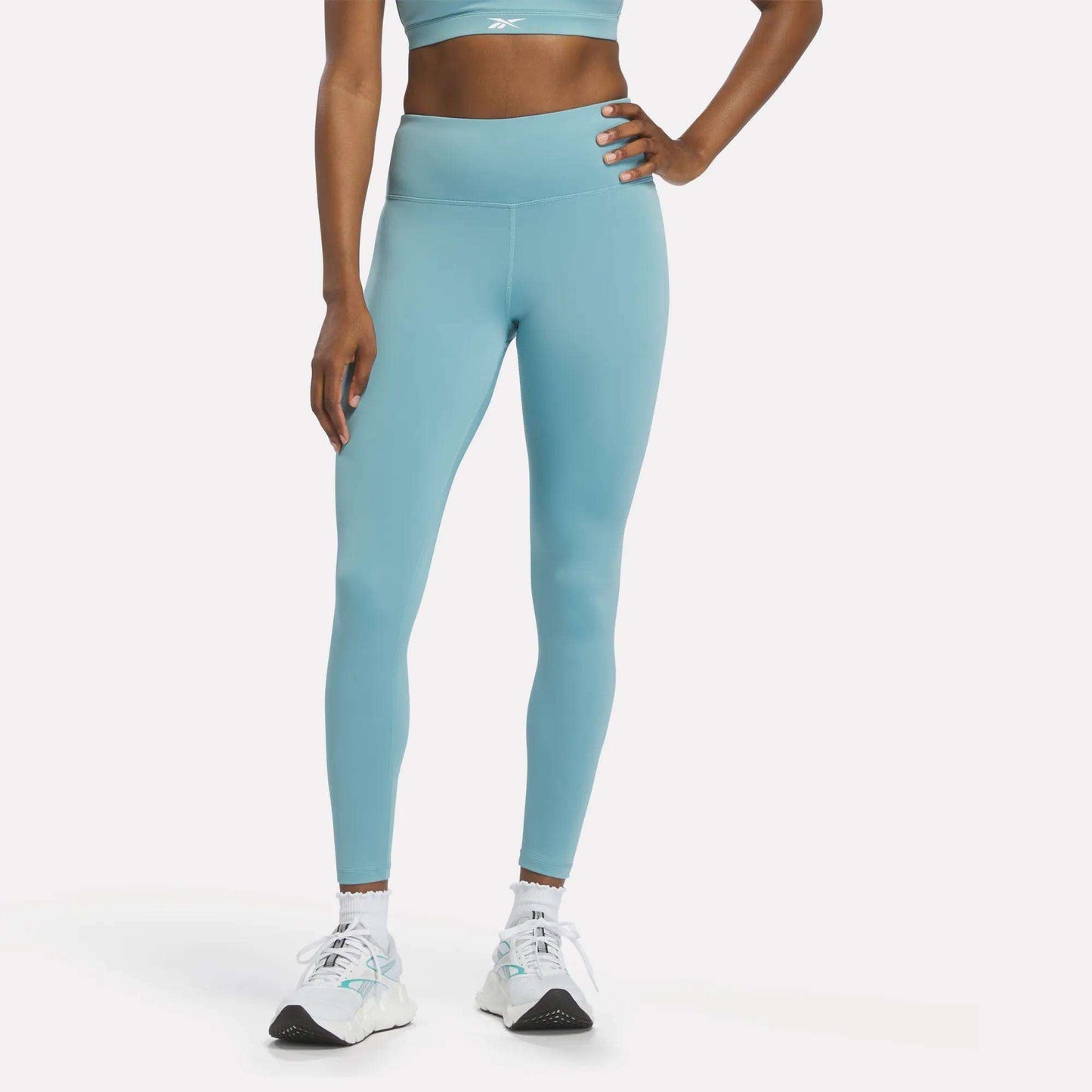 A person wearing Reebok Women's High-Rise Leggings in Hazy Teal, paired with a matching sports bra and white sneakers, stands with one hand on their hip against a plain white background.