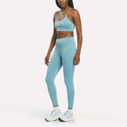 A woman stands facing forward against a plain white background, one arm relaxed by her side. She is wearing the Reebok Women's High-Rise Leggings in light blue with a matching sports bra and white athletic shoes. The leggings are made from comfortable RBK-DRY fabric, designed for performance.