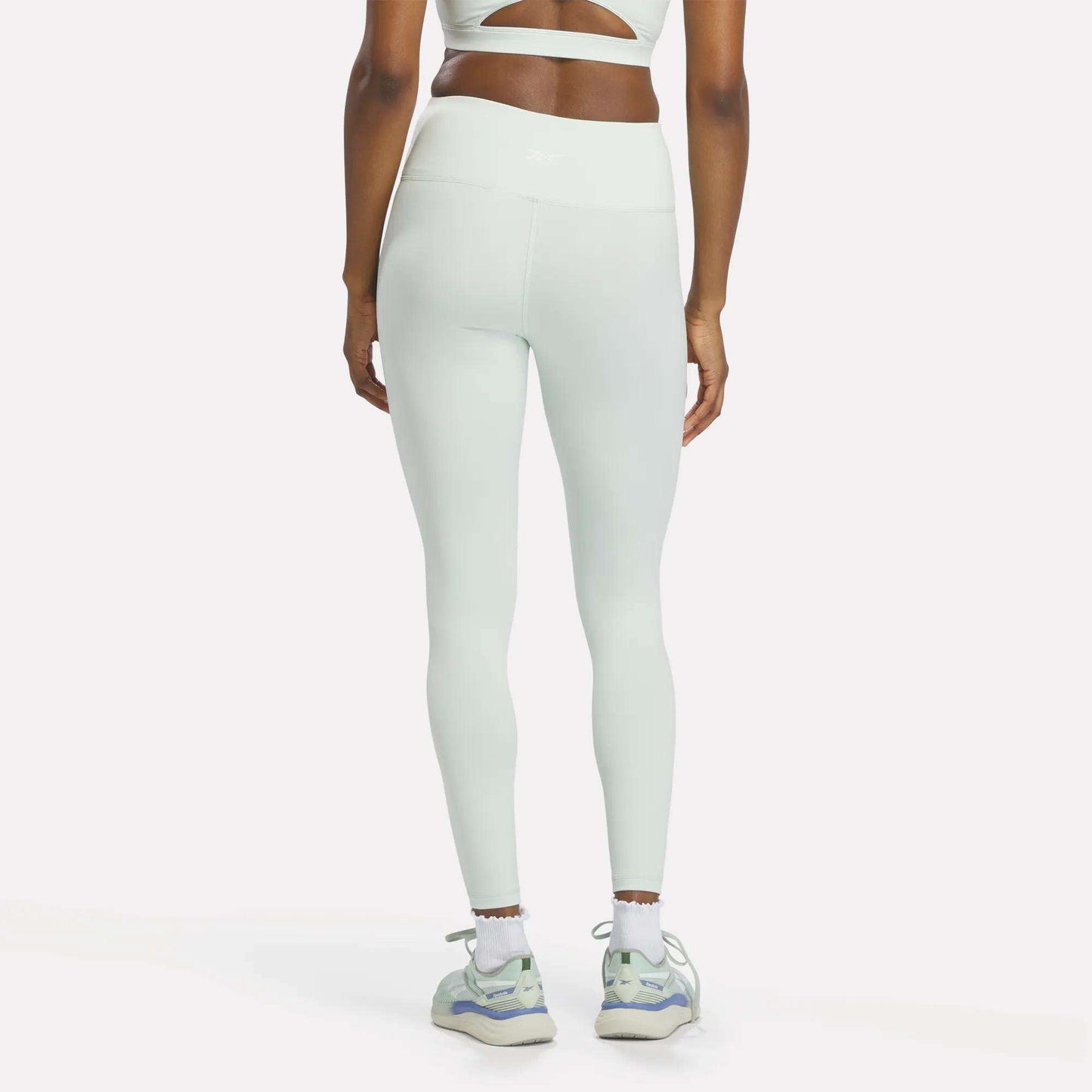 A woman stands facing away, showcasing her back and legs in white Reebok Women's High-Rise Leggings, crafted with RBK-DRY fabric for all-day comfort. She pairs them with a white sports bra, white socks, and white sneakers featuring purple and green accents.