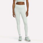 A woman dressed in Reebok Women's High-Rise Leggings in the shade Dew, paired with a matching sports bra, white socks, and mint green sneakers stands against a plain white background. Her head is out of frame.