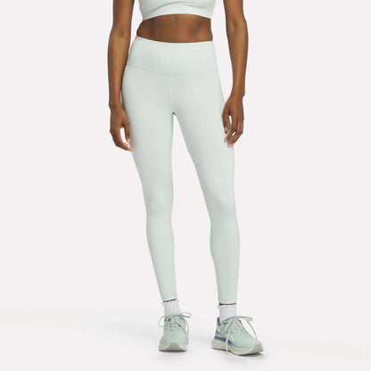 A woman dressed in Reebok Women's High-Rise Leggings in the shade Dew, paired with a matching sports bra, white socks, and mint green sneakers stands against a plain white background. Her head is out of frame.
