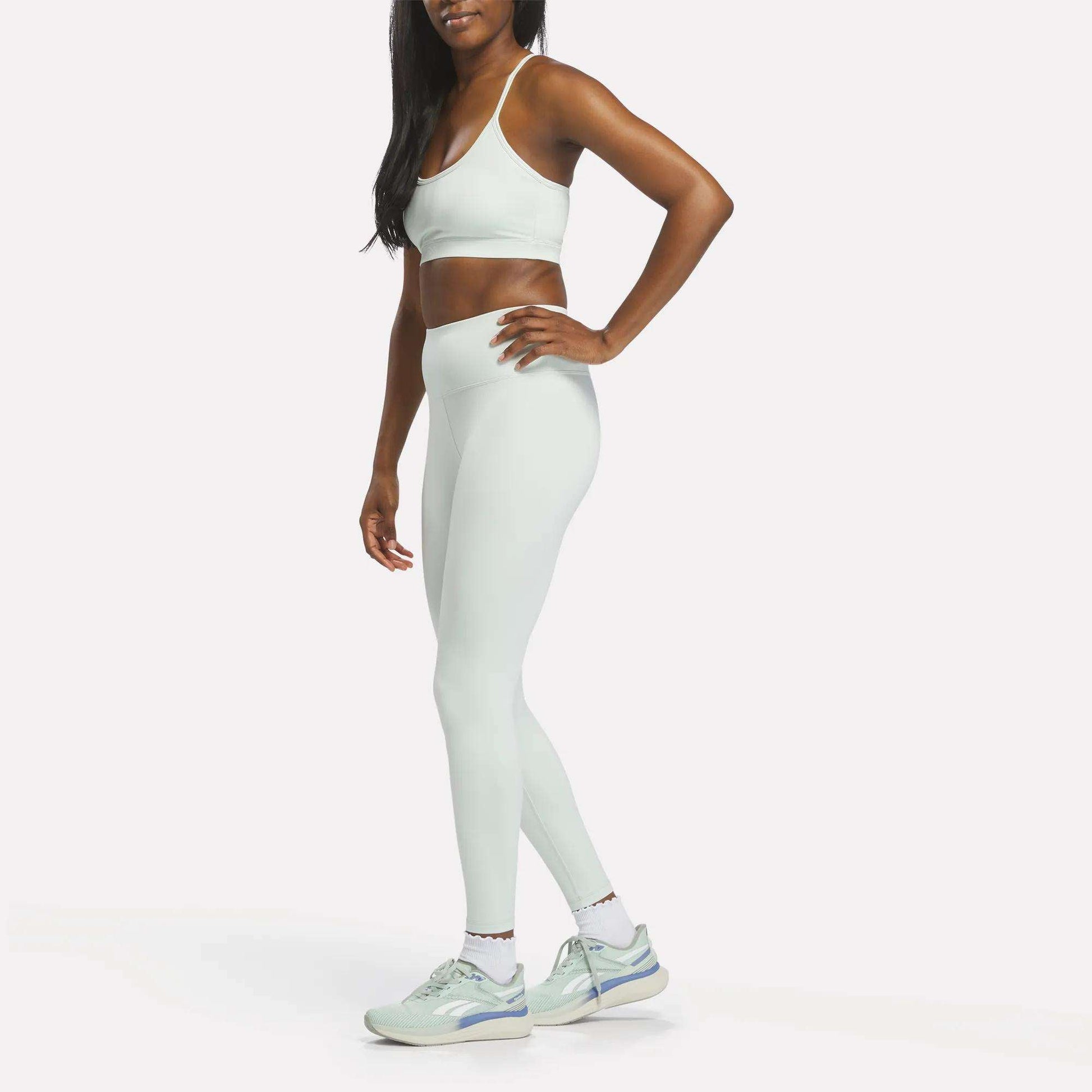 A woman stands in profile wearing Reebok Women's High-Rise Leggings and a matching light blue sports bra, both crafted from moisture-wicking RBK-DRY fabric, along with white sneakers. Her right hand rests on her hip. The background is plain white.