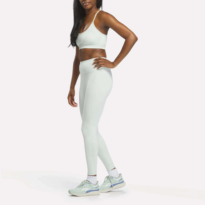 A woman stands in profile wearing Reebok Women's High-Rise Leggings and a matching light blue sports bra, both crafted from moisture-wicking RBK-DRY fabric, along with white sneakers. Her right hand rests on her hip. The background is plain white.