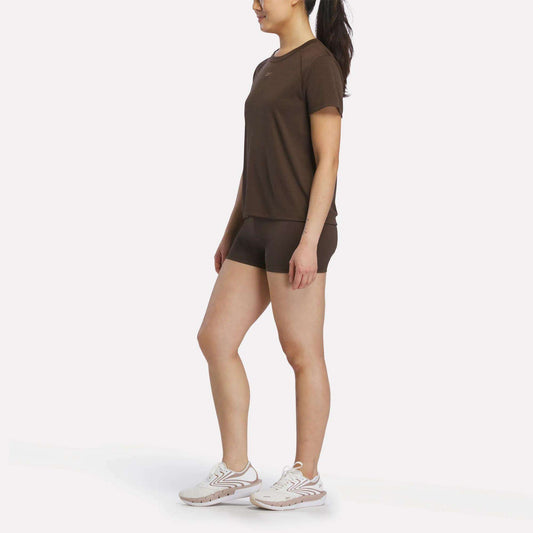 A person stands slightly sideways, wearing a Reebok Women's Training T-Shirt with matching brown shorts and white athletic shoes, set against a plain white background.