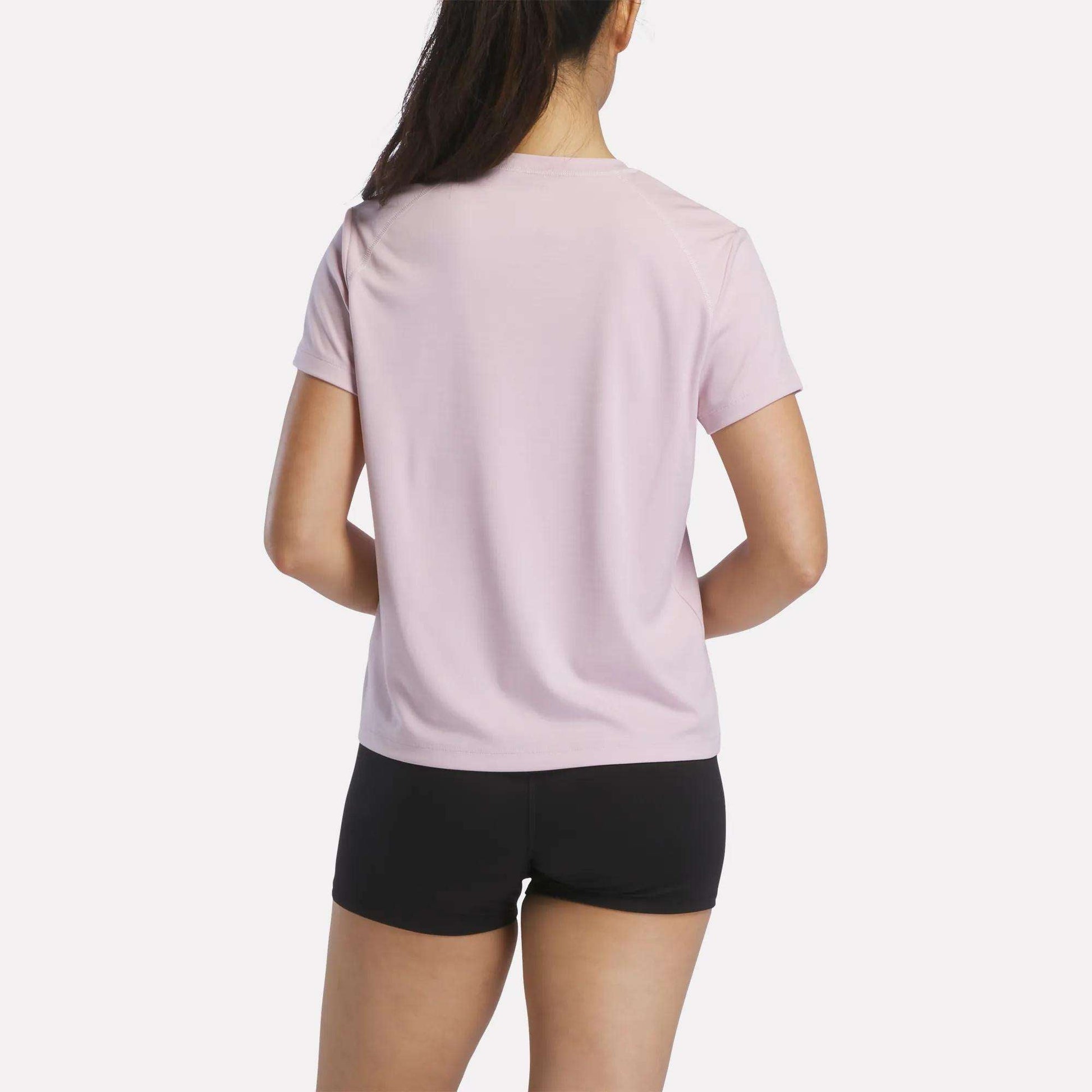 A woman with long dark hair in a ponytail is seen from the back, wearing a light pink Reebok Women's Training T-Shirt and black shorts, standing against a plain white background.