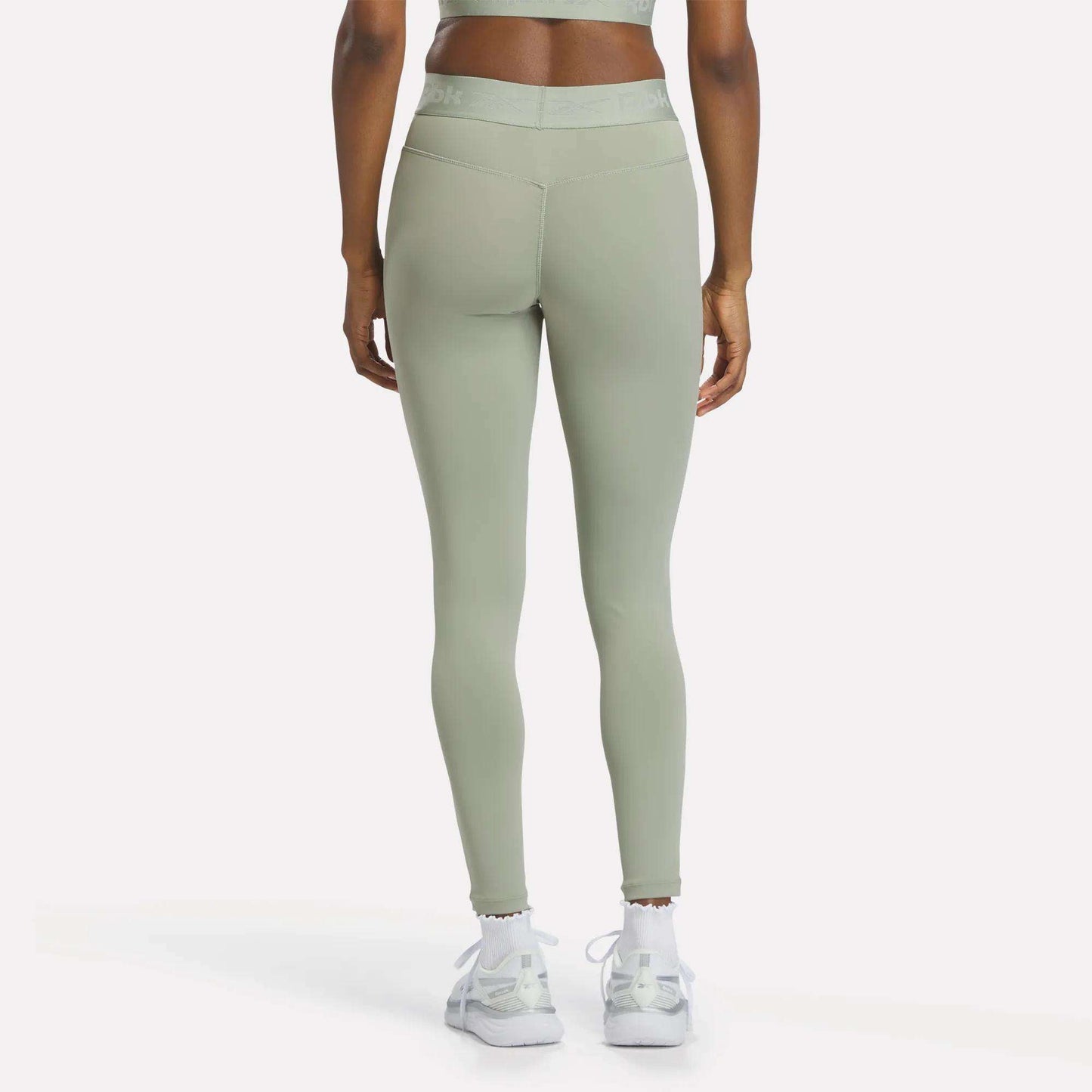 A person wearing Reebok Women's Ruched Training Leggings in light green, made from sweat-wicking fabric, and white sneakers stands facing away from the camera against a plain background.