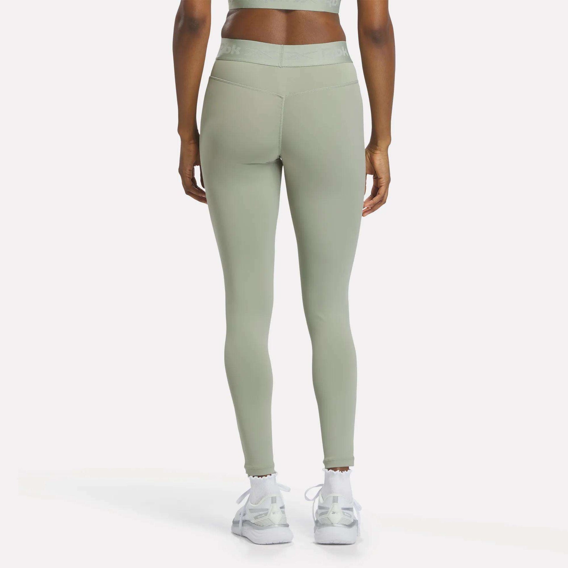 A person wearing Reebok Women's Ruched Training Leggings in light green, made from sweat-wicking fabric, and white sneakers stands facing away from the camera against a plain background.