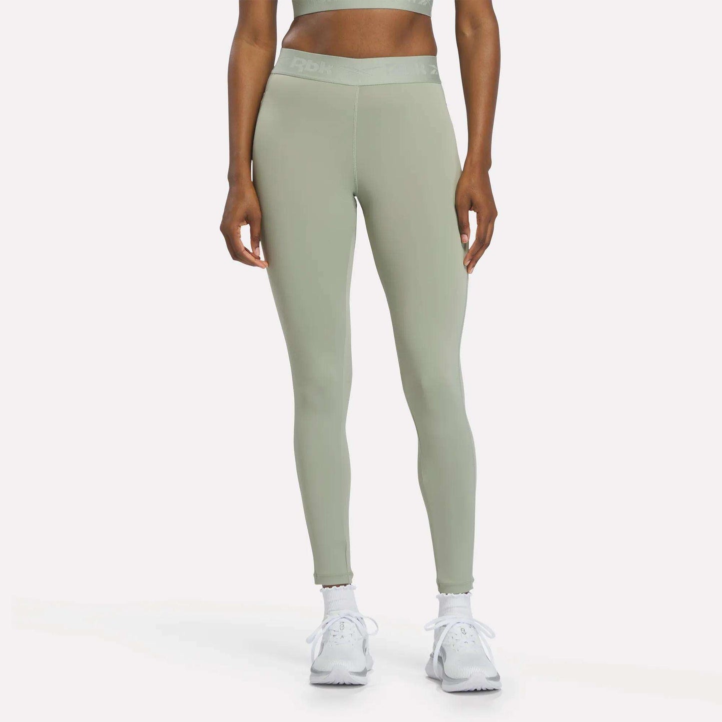 A person is shown from the waist down, standing against a plain white background, wearing Reebok Women's Ruched Training Leggings in Hazy Green, made from sweat-wicking fabric, paired with white sneakers.