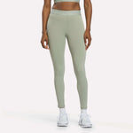 A person is shown from the waist down, standing against a plain white background, wearing Reebok Women's Ruched Training Leggings in Hazy Green, made from sweat-wicking fabric, paired with white sneakers.