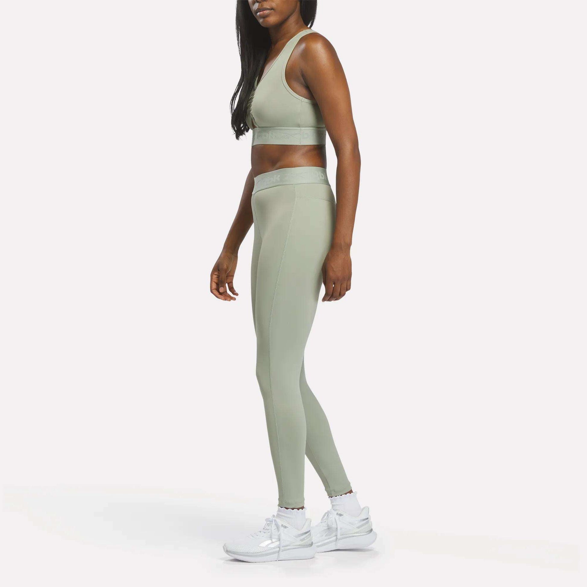 A woman stands sideways against a plain white background, looking forward, dressed in sage green Reebok Women's Ruched Training Leggings paired with a matching sports bra and white sneakers. Her outfit is crafted from sweat-wicking fabric for optimal comfort.