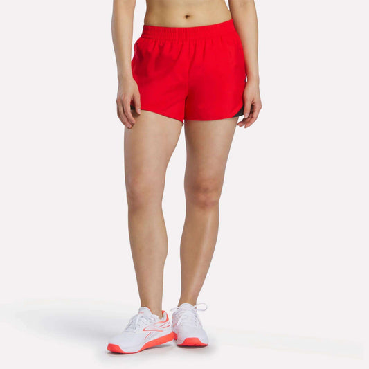 A person stands against a plain background wearing Reebok Women's Colorblocked Woven Shorts in Vector Red with an elastic waistband and white sneakers featuring red accents. Only their lower body is visible.