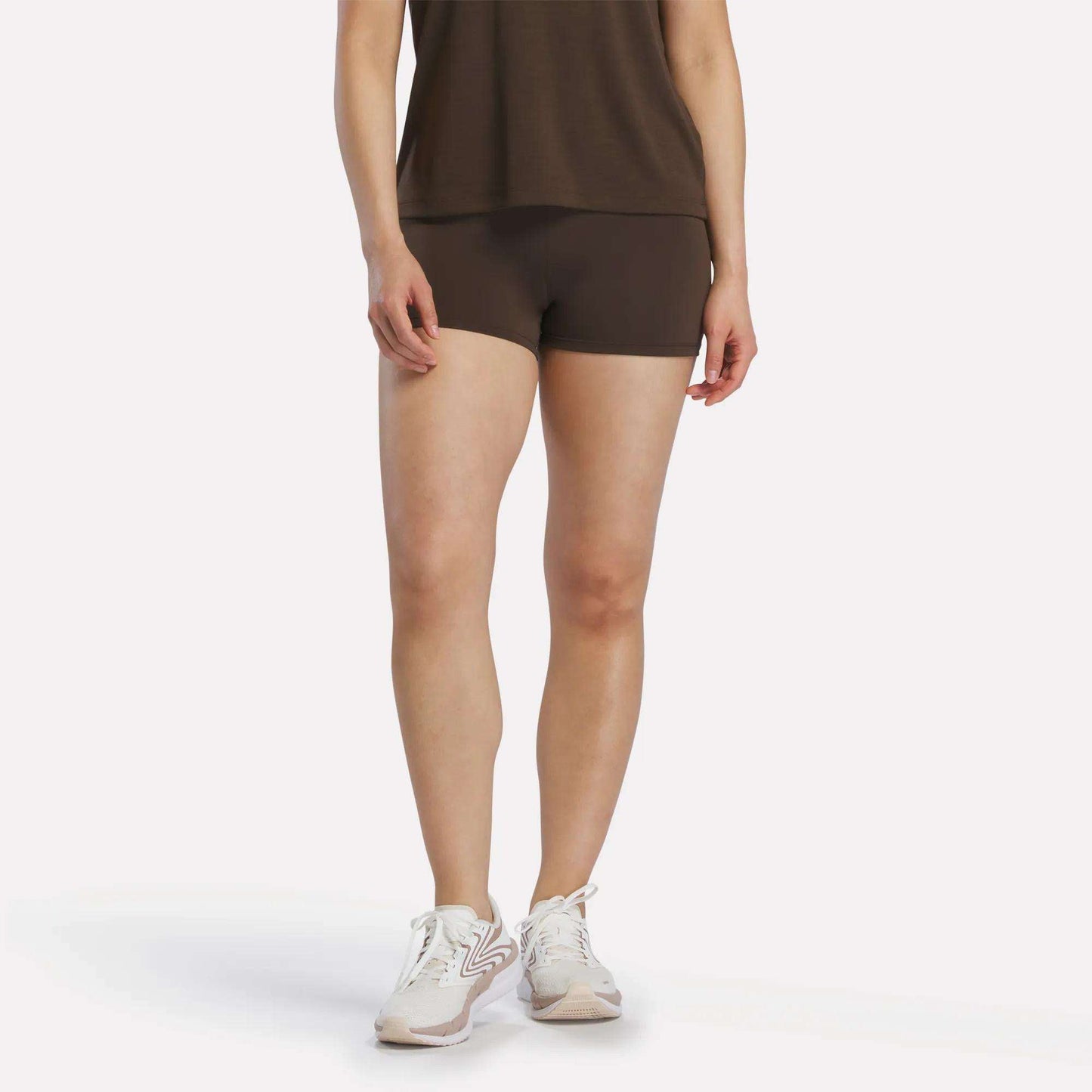 A person stands against a plain white background, wearing an espresso-colored Reebok Women's High-Rise Mini Biker Shorts 3" paired with a brown top and white sneakers. Their head is not visible in the image.