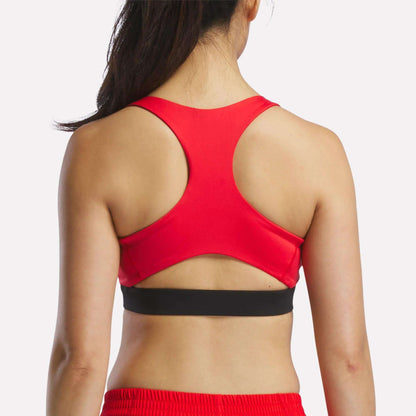 A woman, seen from behind, wears a red Reebok Women's Racer Sports Bra featuring a racerback design for mid-impact support, paired with matching red shorts. Her hair is styled in a ponytail, and the background is plain white.