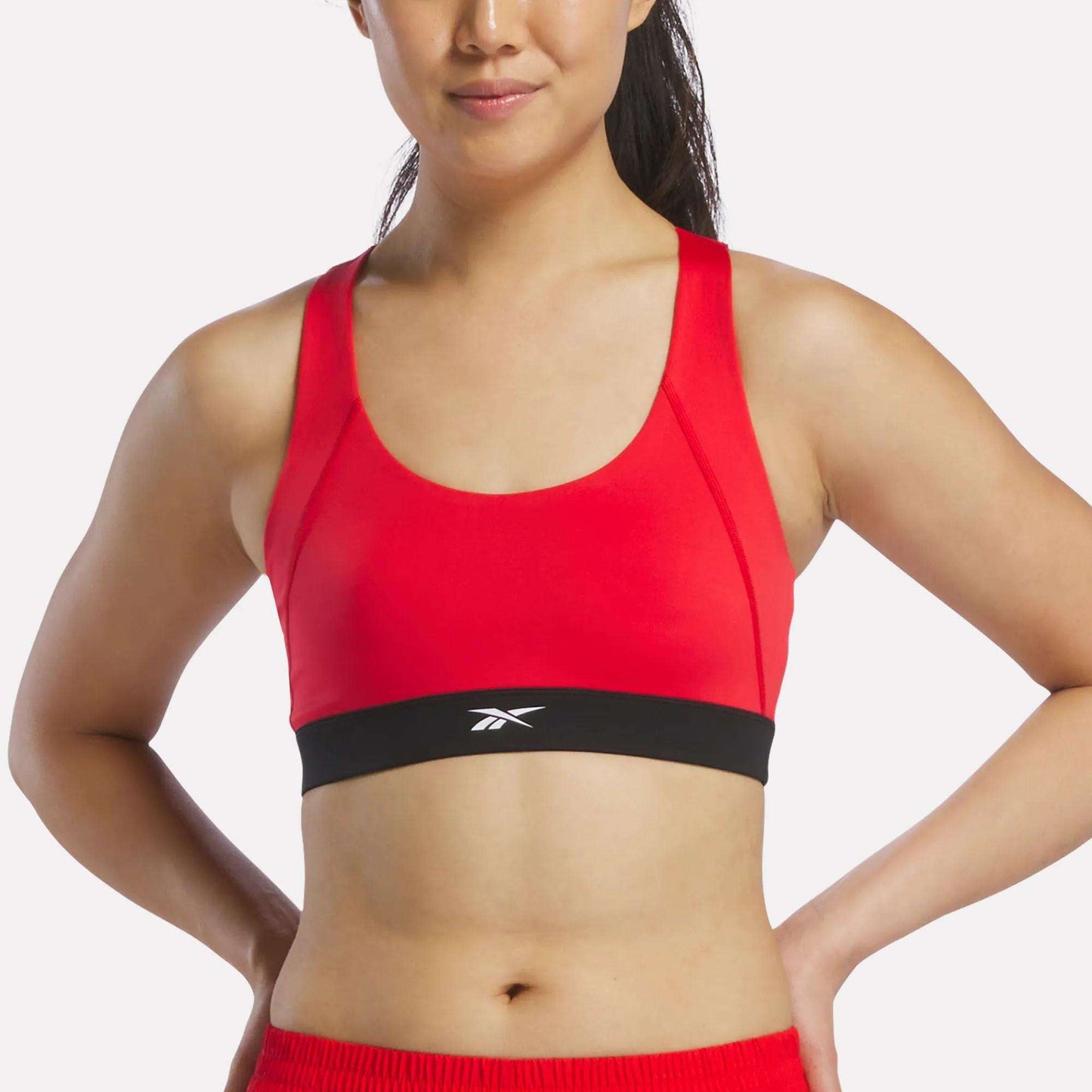 A woman stands with her hands on her hips against a plain background, wearing the Reebok Women's Racer Sports Bra in Vector Red, which features a black band with a white logo. This stylish bra provides mid-impact support, making it ideal for active routines.