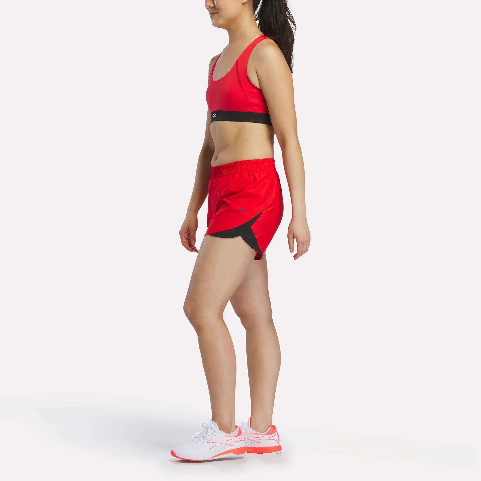 A woman in a red Reebok Women's Racer Sports Bra, providing mid-impact support, paired with red shorts featuring black accents and white athletic shoes, stands in a side-facing pose against a plain white background.