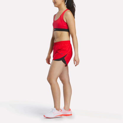 A woman in a red Reebok Women's Racer Sports Bra, providing mid-impact support, paired with red shorts featuring black accents and white athletic shoes, stands in a side-facing pose against a plain white background.