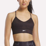 A woman wearing the Reebok Women's Printed Tri-Back Sports Bra in black, featuring thin straps and the Reebok logo on the front, stands against a plain white background.