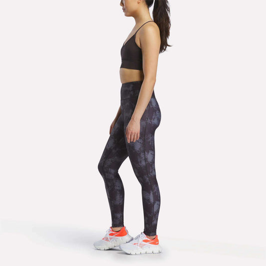 A woman wearing athletic apparel—including the Reebok Women's Printed Tri-Back Sports Bra, patterned leggings, and white sneakers—standing in profile against a plain white background.