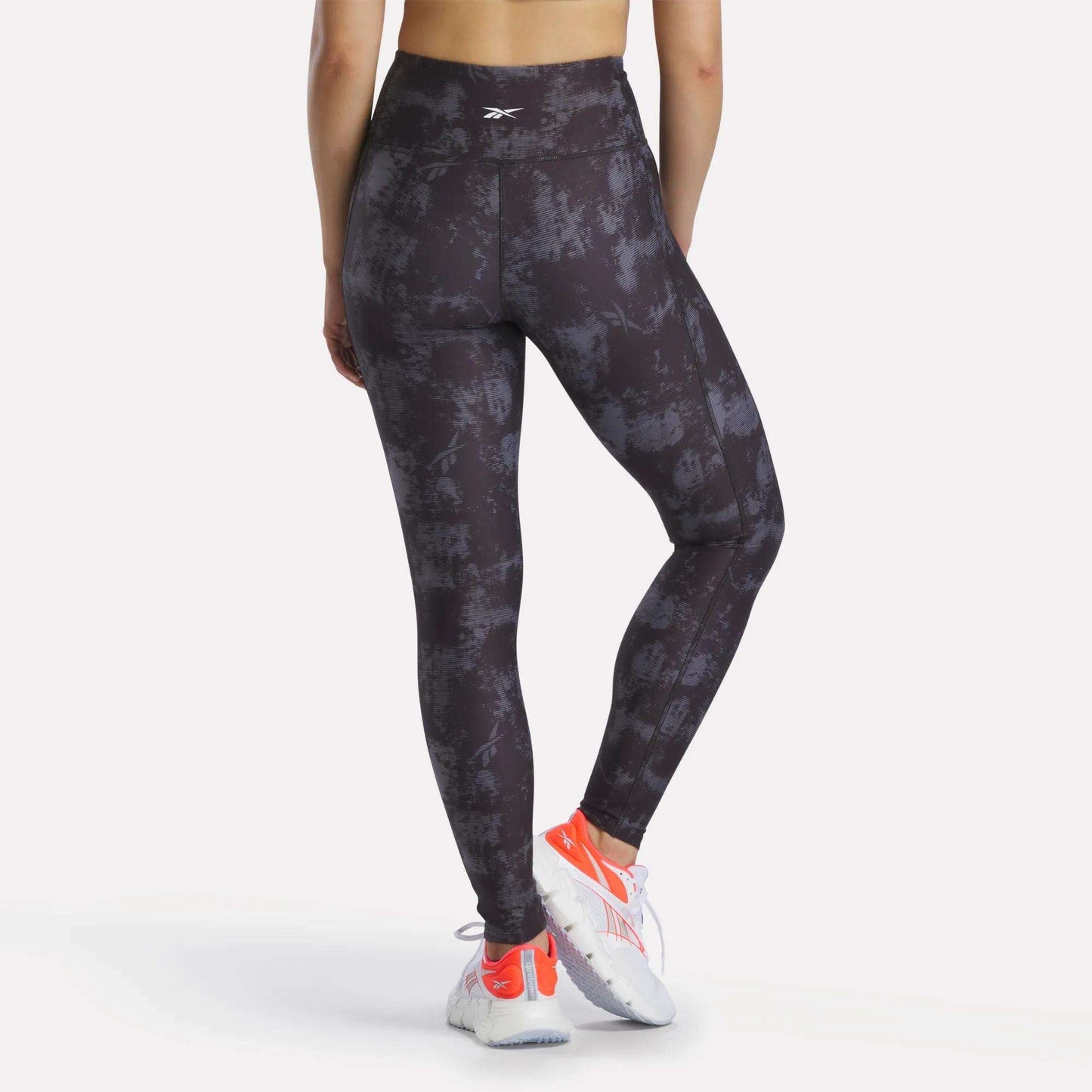 A person faces away, wearing Reebok Women's High-Rise Printed Leggings with sweat-wicking fabric, white and orange athletic shoes, and a sports top. The background is plain and light-colored.