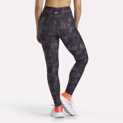 A person faces away, wearing Reebok Women's High-Rise Printed Leggings with sweat-wicking fabric, white and orange athletic shoes, and a sports top. The background is plain and light-colored.