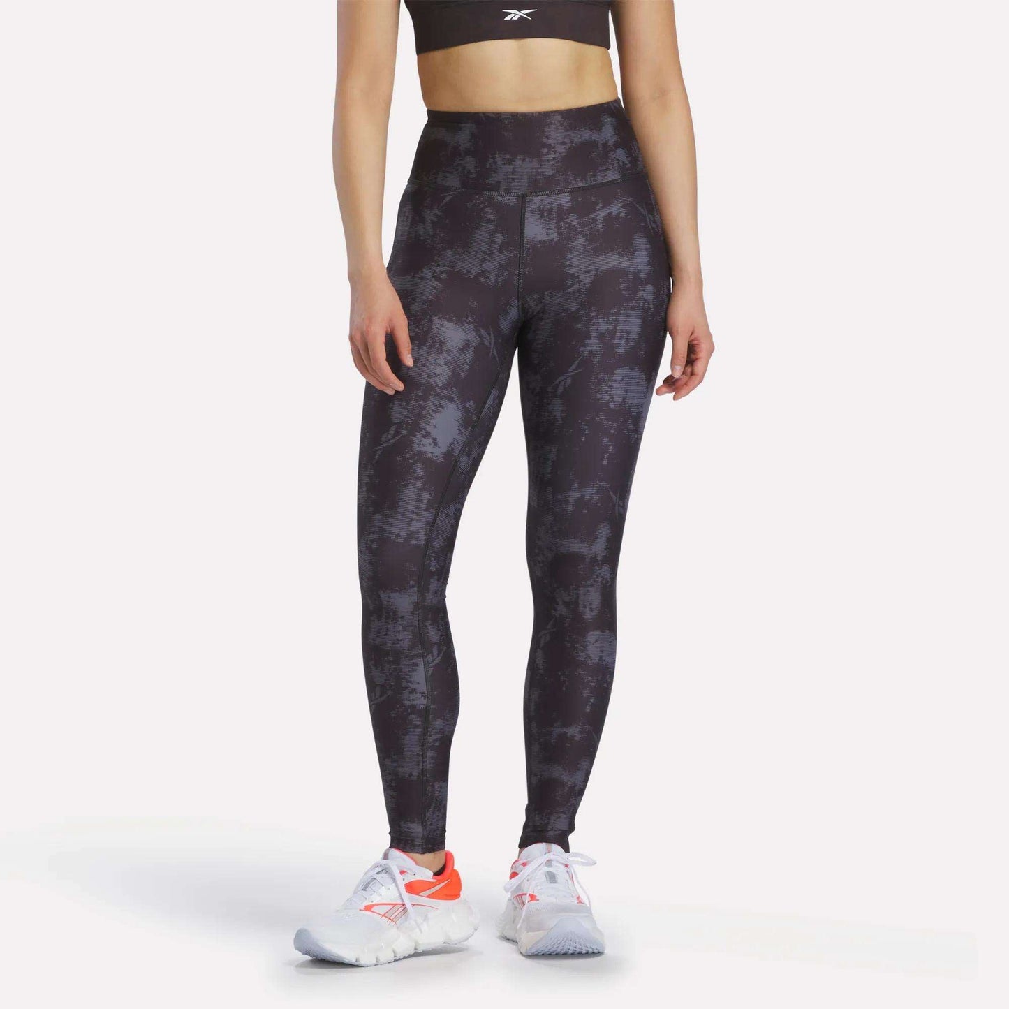 A woman in a black sports bra, Reebok Women's High-Rise Printed Leggings with sweat-wicking fabric, and white athletic shoes with orange accents stands against a plain light background.
