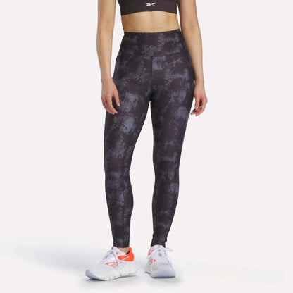 A woman in a black sports bra, Reebok Women's High-Rise Printed Leggings with sweat-wicking fabric, and white athletic shoes with orange accents stands against a plain light background.